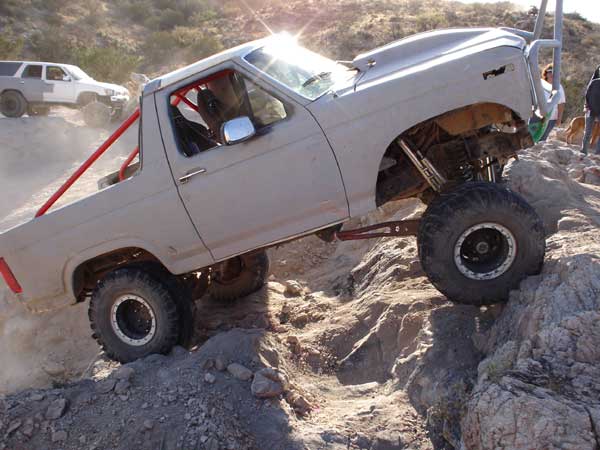 Gray off-road truck with red roll cage climbing rocks, another white SUV in background