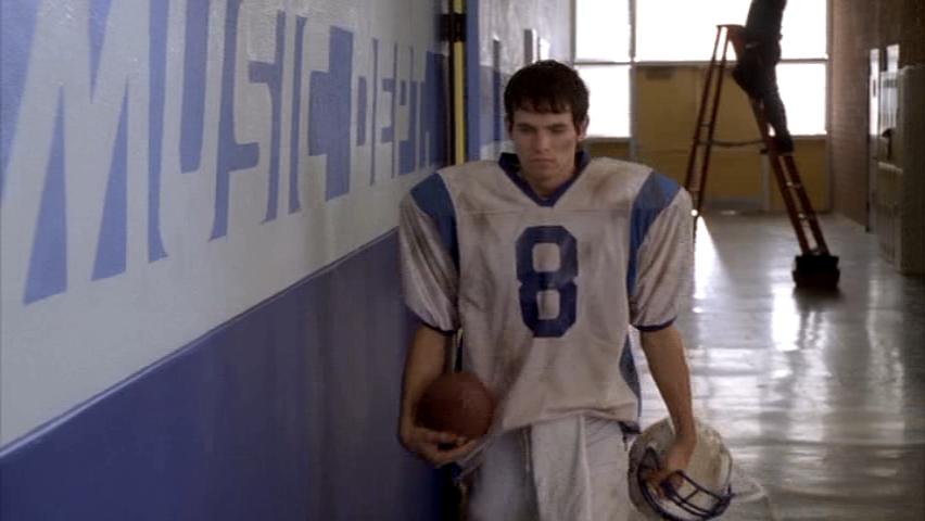 Football player with jersey number 8 walks down a school hallway holding a football and helmet. Text is on the wall