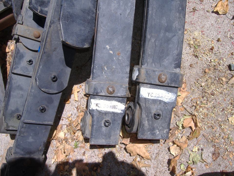Dark metal leaf spring suspension parts with labeled stickers lying on gravel ground.