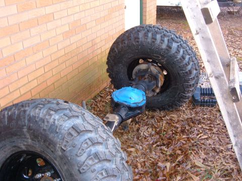 1987 Ford Bronco - Vehicle axle and tires are shown resting on the ground next to a brick wall.