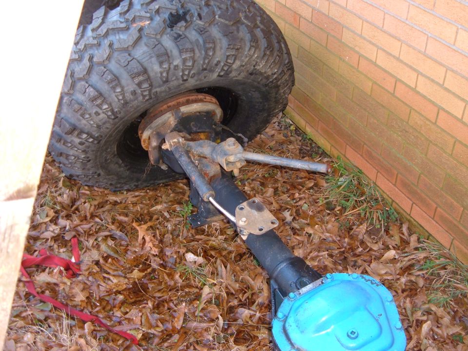 Vehicle axle with large tire, sitting on the ground near a brick wall