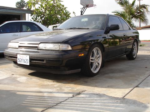 1989 Mazda MX-6 - This black 1989 Mazda MX-6 coupe is parked in a driveway with another white car visible.