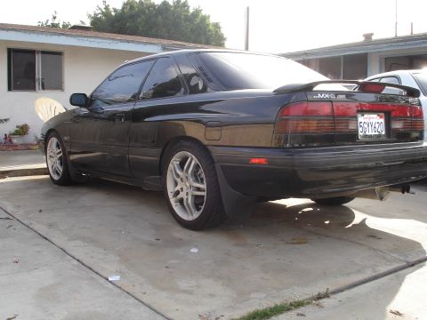1989 Mazda MX-6 - A black 1989 Mazda MX-6 is parked in a driveway, showing its rear profile and custom wheels.