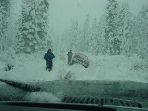 1986 Ford Bronco - A red SUV is stuck in deep snow in a wooded area, with two people observing the scene.