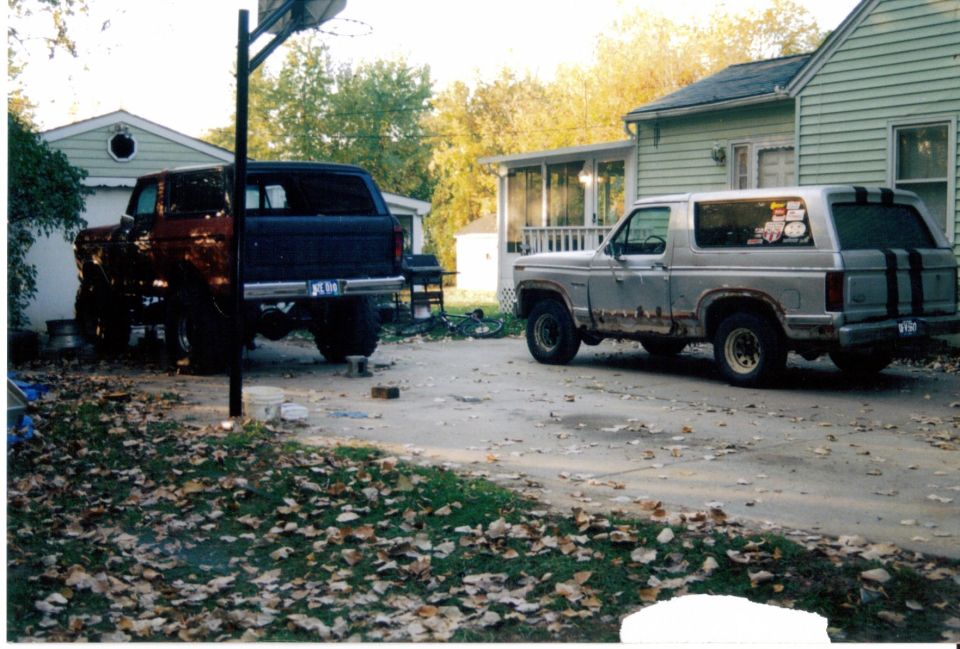 Two Ford Broncos, one dark with rust-red panels and one silver with black stripes, parked in a leaf-strewn driveway