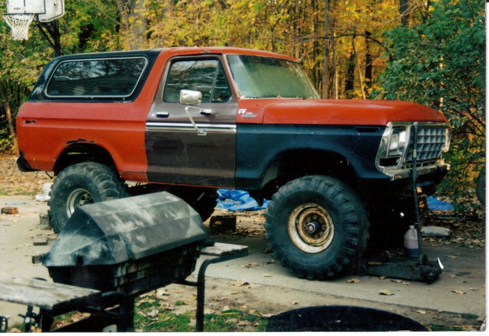 Lifted red Ford Bronco with primer panels and big off-road tires parked in a leafy driveway, grill in foreground
