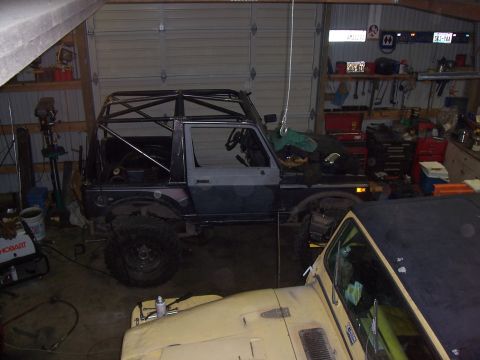 1988 Suzuki Samurai - A heavily modified off-road vehicle with an extensive roll cage is seen in a garage setting, partially obscured by another vehicle.
