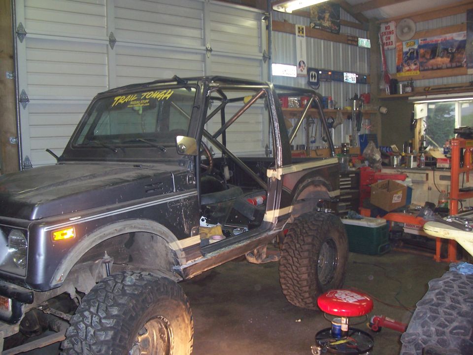 Dark Suzuki Samurai with roll cage and "Trail Tough" sticker, in a garage