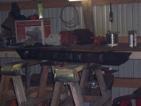 1988 Suzuki Samurai - A black metal bar with multiple circular cutouts is supported by wooden sawhorses in a cluttered workshop setting.
