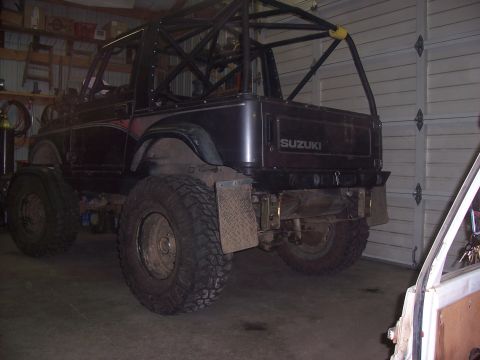 1988 Suzuki Samurai - A dark Suzuki Samurai with a custom roll cage and oversized tires is parked indoors.