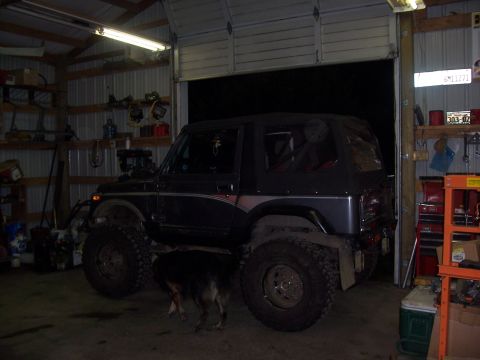 1988 Suzuki Samurai - A modified dark SUV with oversized tires and a soft top is parked inside a cluttered garage.