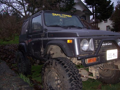 1988 Suzuki Samurai - A dark grey Suzuki Samurai with off-road modifications sits on uneven terrain.