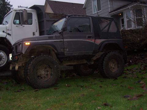 1988 Suzuki Samurai - A dark gray off-road vehicle with a soft top is parked on a grassy area next to a white truck.