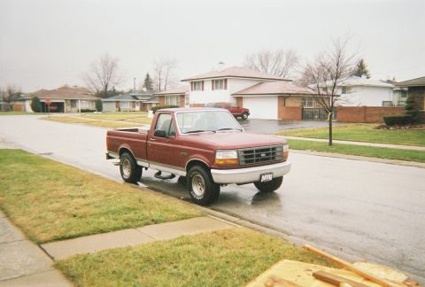 1996 Ford F-150 - A red pickup truck is parked on the side of a wet street in a residential neighborhood.