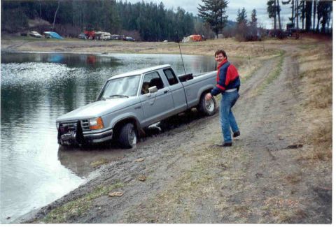 1989 Ford Bronco - A man watches as his Ford Ranger pickup truck gets stuck in the water near a lake.