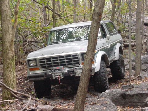 1978 Ford Bronco - A 1978 Ford Bronco is shown off-roading through a dense forest with large rocks and mud.