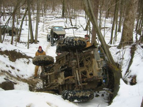 1992 Ford Bronco - An off-road vehicle has overturned in a snowy, wooded area, with two people observing the scene.