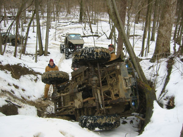 off-road vehicle flipped on its side in a snowy wooded area with two people nearby and another vehicle in the background