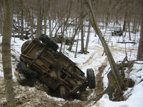 1992 Ford Bronco - An overturned off-road vehicle lies in a muddy, snowy ditch surrounded by trees, with another vehicle visible in the distance.