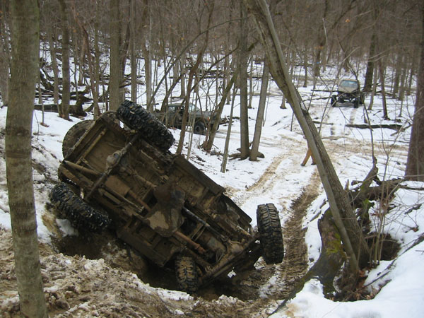 Mud-covered off-road vehicle overturned in a snowy ditch, trees and another vehicle in background