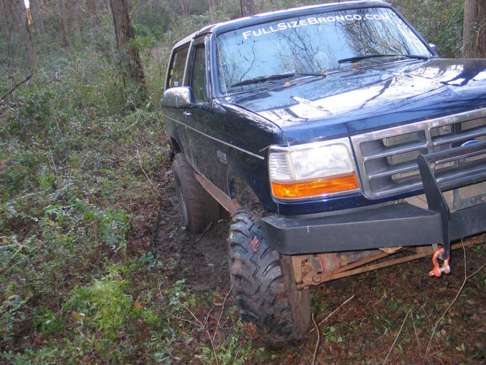 Blue 1995 Ford Bronco with "FULLSIZEBRONCO.COM" sticker on windshield, stuck in mud on wooded trail