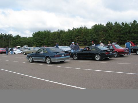 1990 Ford Mustang - Two Ford Mustangs, a blue and a black model, are lined up on a racetrack with other vehicles in the background.