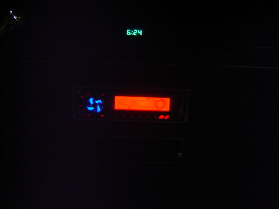 Car stereo with orange display and blue fan, above it a green digital clock reads 6:24
