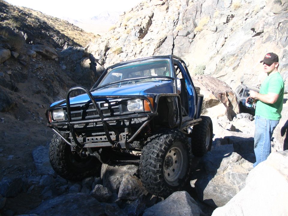 Blue Toyota pickup with tube bumper and big tires on rocks, man in green shirt spotting
