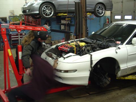 1996 Ford Probe - Mechanics work on removing the engine from a white car in a garage. Another car is lifted on a hydraulic lift above.