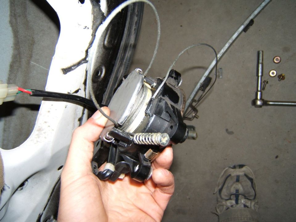 Hand holding a car window regulator mechanism with cables and wires attached. Tools and hardware are on the floor