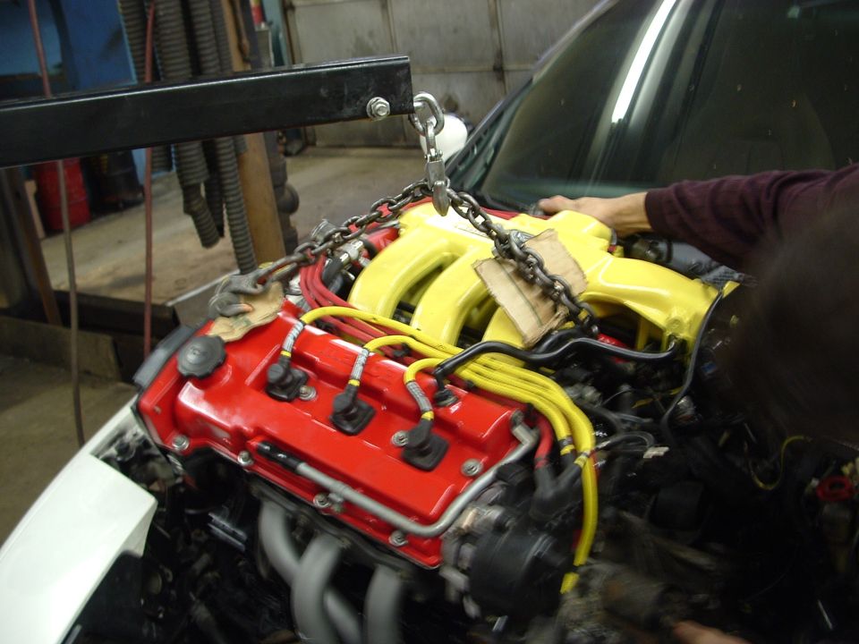 Engine with red valve cover and yellow intake manifold being lifted by a chain hoist