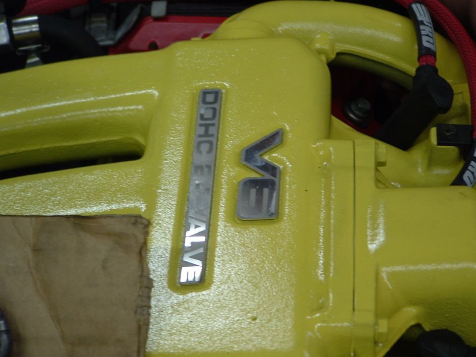 Close-up of a yellow engine component with "DOHC 24 VALVE" and "V6" emblems. Some brown cardboard is visible on the left