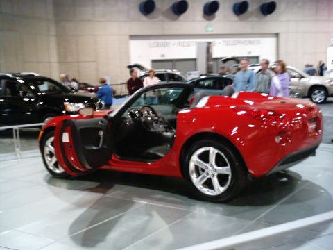 1986 Ford Bronco - A red convertible sports car is displayed indoors with its door open, alongside other vehicles.