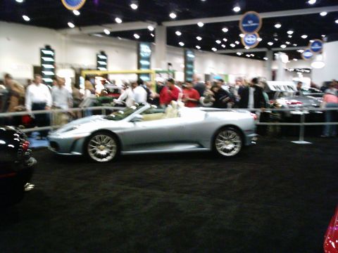 1986 Ford Bronco - A silver convertible sports car is shown at an auto show with people in the background.