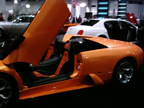 1986 Ford Bronco - An orange mid-engine exotic coupe on an indoor auto-show floor with its scissor door raised, showing orange-and-black leather seats and doo…
