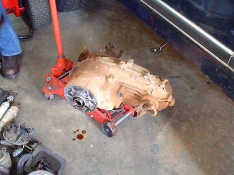 1984 Ford Bronco - A transmission assembly is supported by a red jack, with various automotive parts scattered around the garage floor.