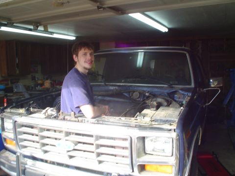 1984 Ford Bronco - A man is seen working on the engine of a blue pickup truck in a garage.