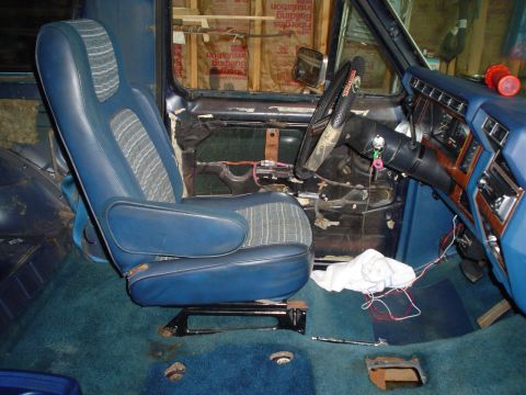 1984 Ford Bronco - The interior of a vehicle shows a blue seat, dashboard, and steering wheel, with exposed wiring and unfinished walls.