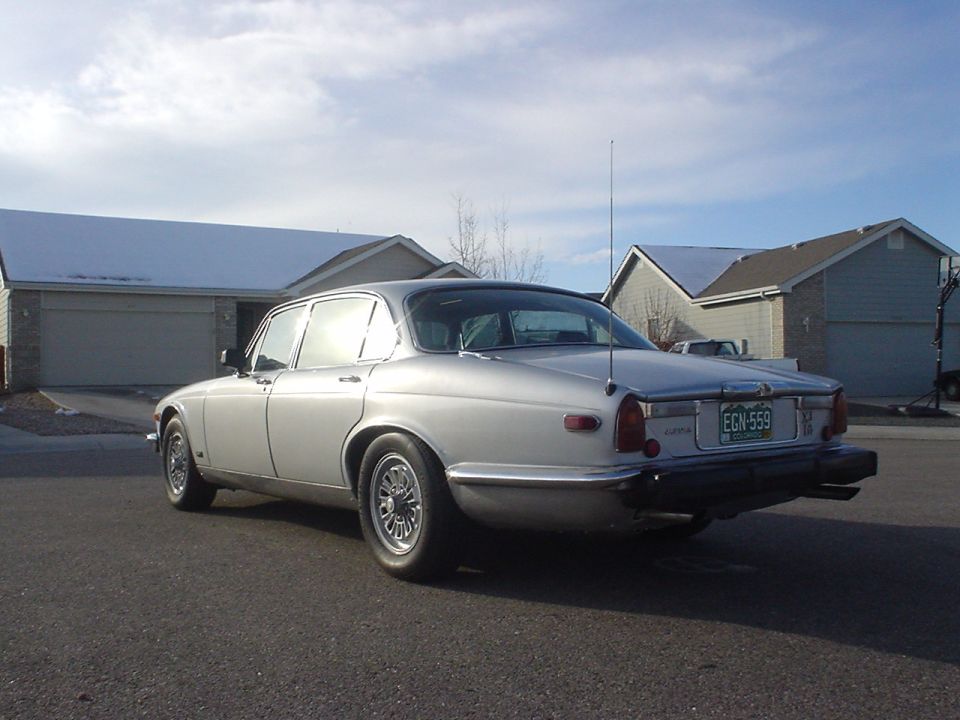 Rear three-quarter view of a silver Jaguar XJ6 sedan parked on a street in front of suburban houses
