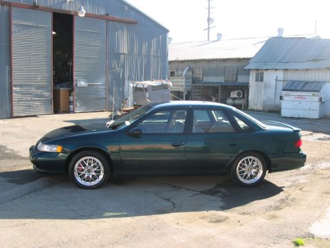 1995 Ford Taurus - A dark green 1995 Ford Taurus SHO sedan is parked outside a corrugated metal building, its side profile and aftermarket wheels.