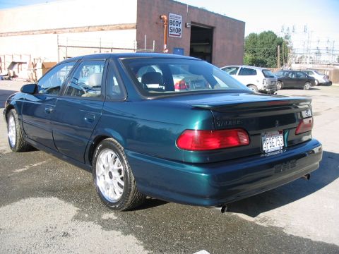 1995 Ford Taurus - A 1995 Ford Taurus sedan is seen from the rear outside a body shop with other vehicles in the background.