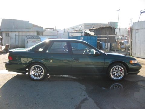 1995 Ford Taurus - Side profile of a dark green 1995 Ford Taurus sedan with aftermarket wheels parked outdoors.
