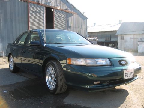 1995 Ford Taurus - A dark green sedan is parked outside a metal building on a sunny day.