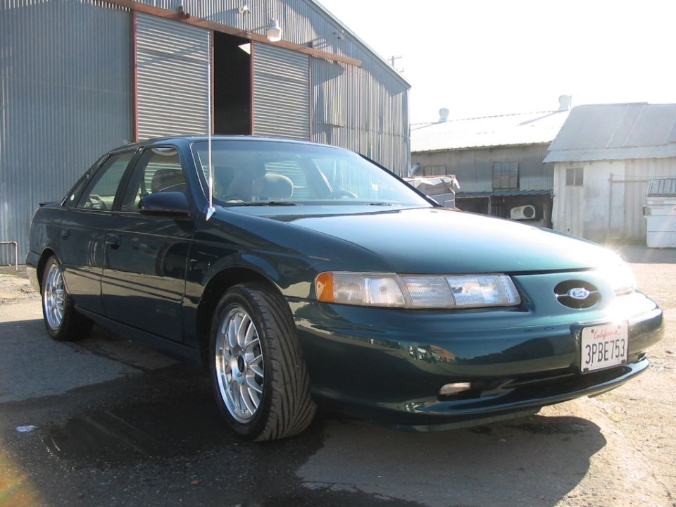 Dark green sedan parked in front of a corrugated metal building. The car has aftermarket wheels and a California license plate reading 3PBE753
