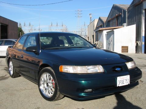 1995 Ford Taurus - A dark green sedan is parked in front of industrial buildings under a clear blue sky.