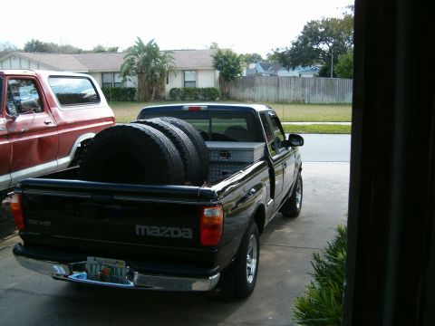 1979 Ford Bronco - A black Mazda pickup truck is parked next to a red Ford Bronco, with tires loaded in the truck bed.