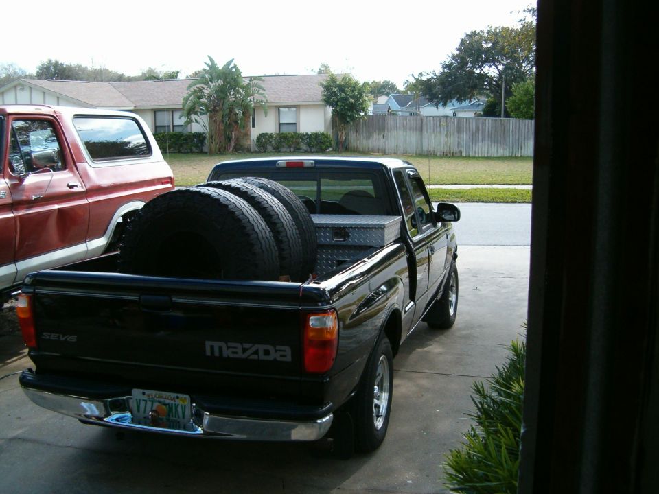 Black Mazda pickup truck rear view, carrying three tires and a toolbox
