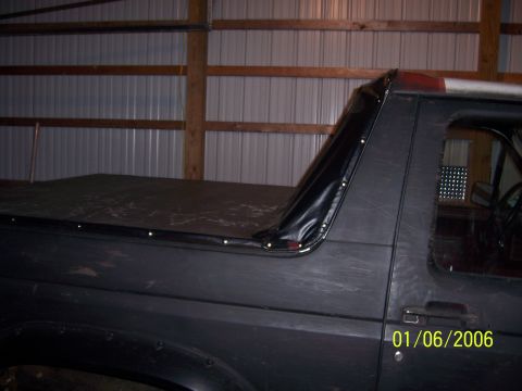 1985 Ford Bronco - The side of a Ford Bronco is shown with a black soft top covering the rear cargo area. The vehicle is parked indoors.