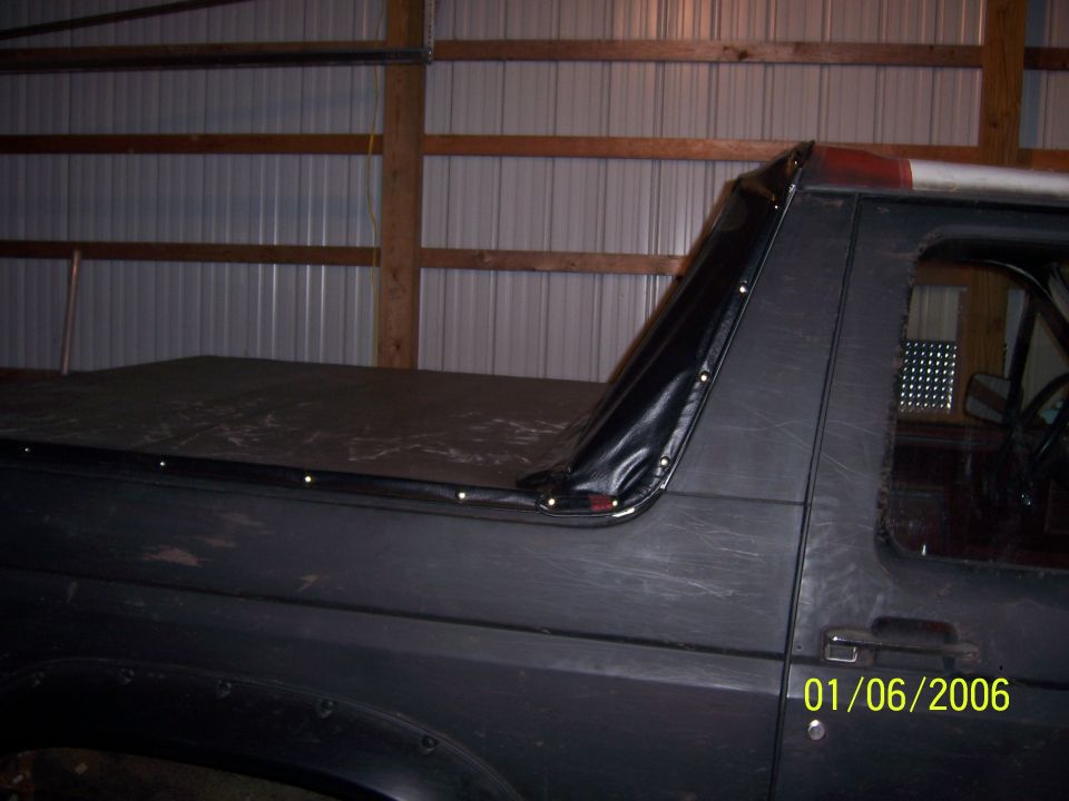 Side view of a dark-colored Ford Bronco with a black soft top installed over the bed. The vehicle is inside a barn