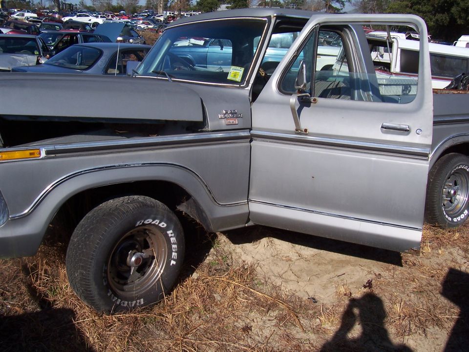 Silver Ford F-Series pickup truck with door open, parked in a junkyard with other vehicles